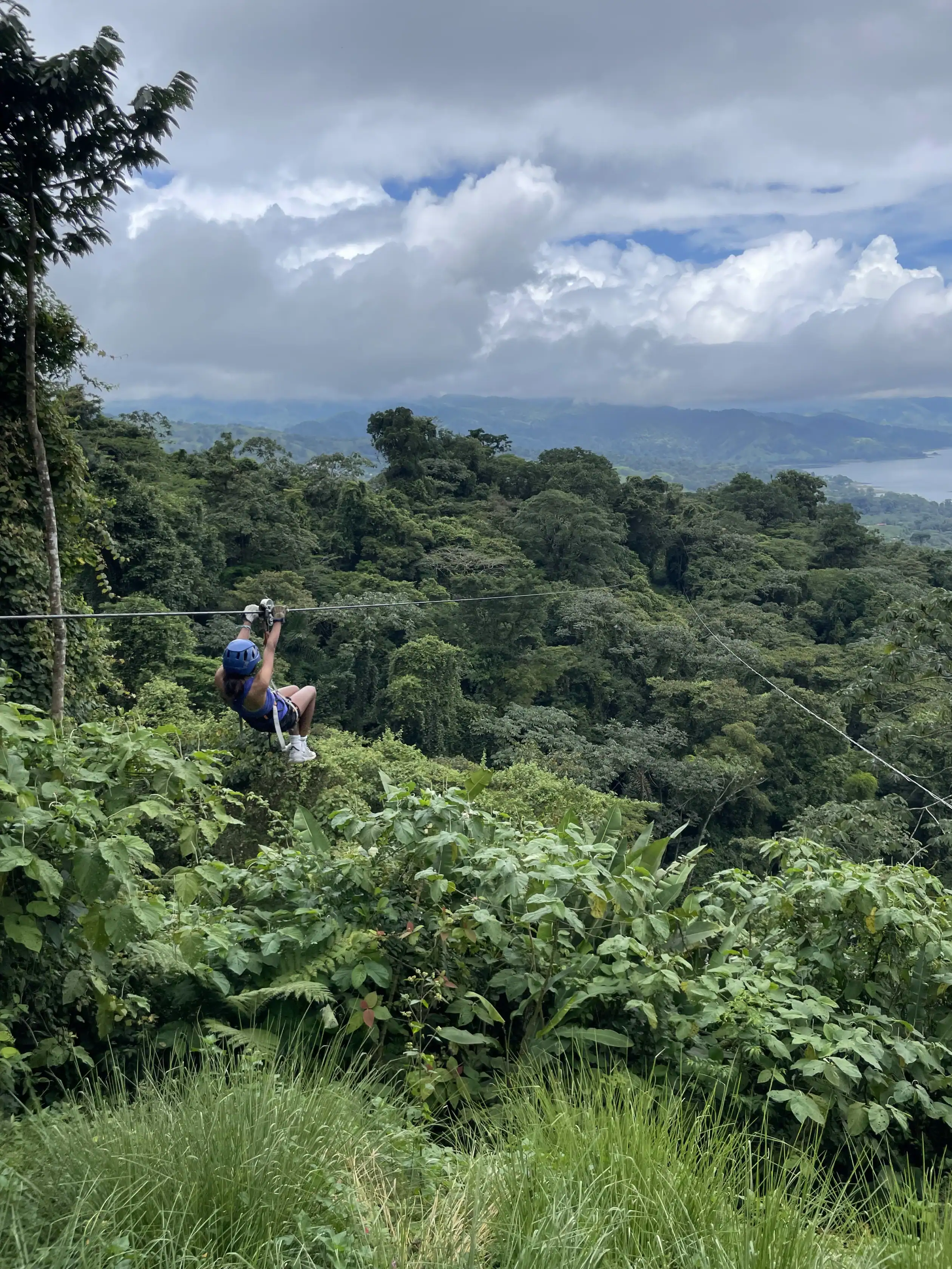 A person wearing a helmet and harness ziplines over lush, green forest with hills and a cloudy sky in the background on a teen medical trip abroad.