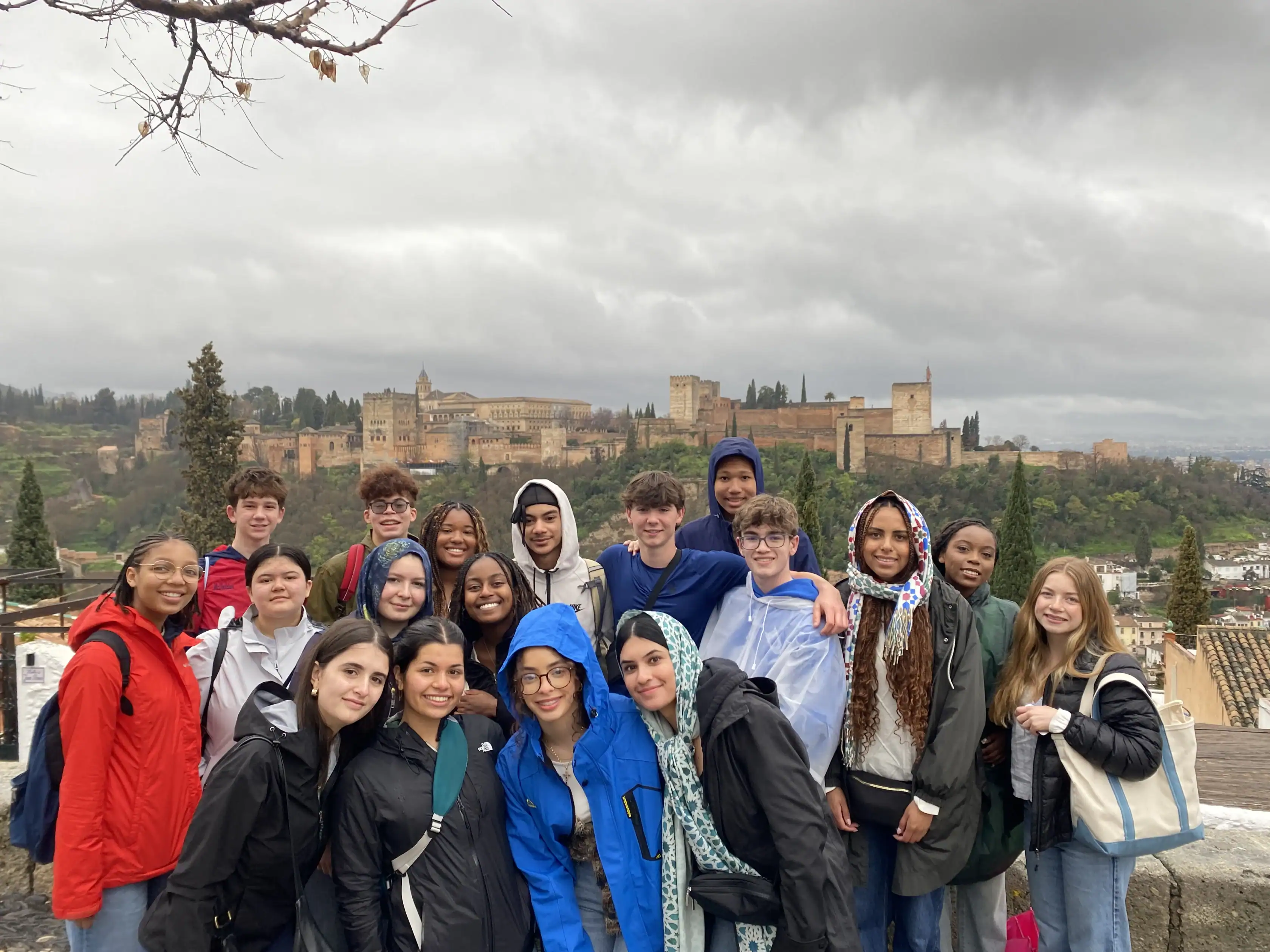 A group of teenagers pose together outdoors on a cloudy day with a historic fortress and greenery visible in the background.