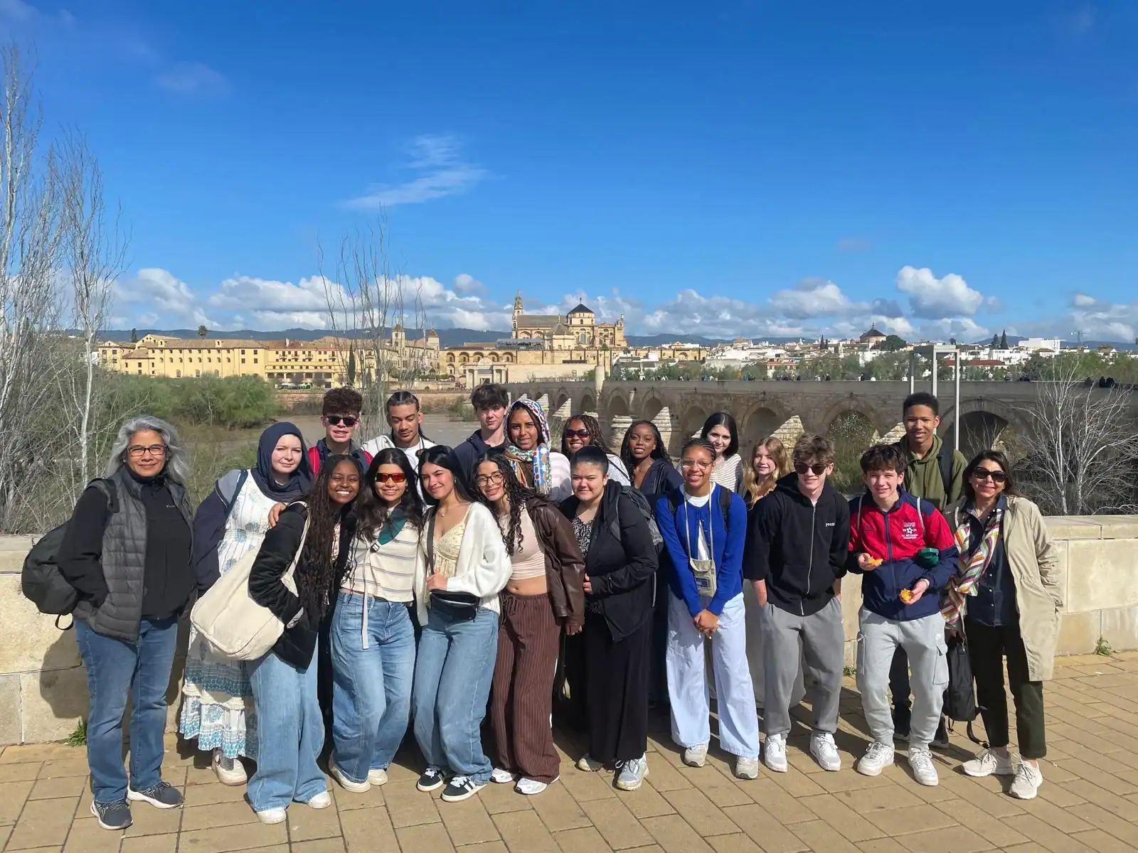 A group of people pose for a photo outdoors in front of a stone railing, with a historic cityscape and blue sky in the background.
