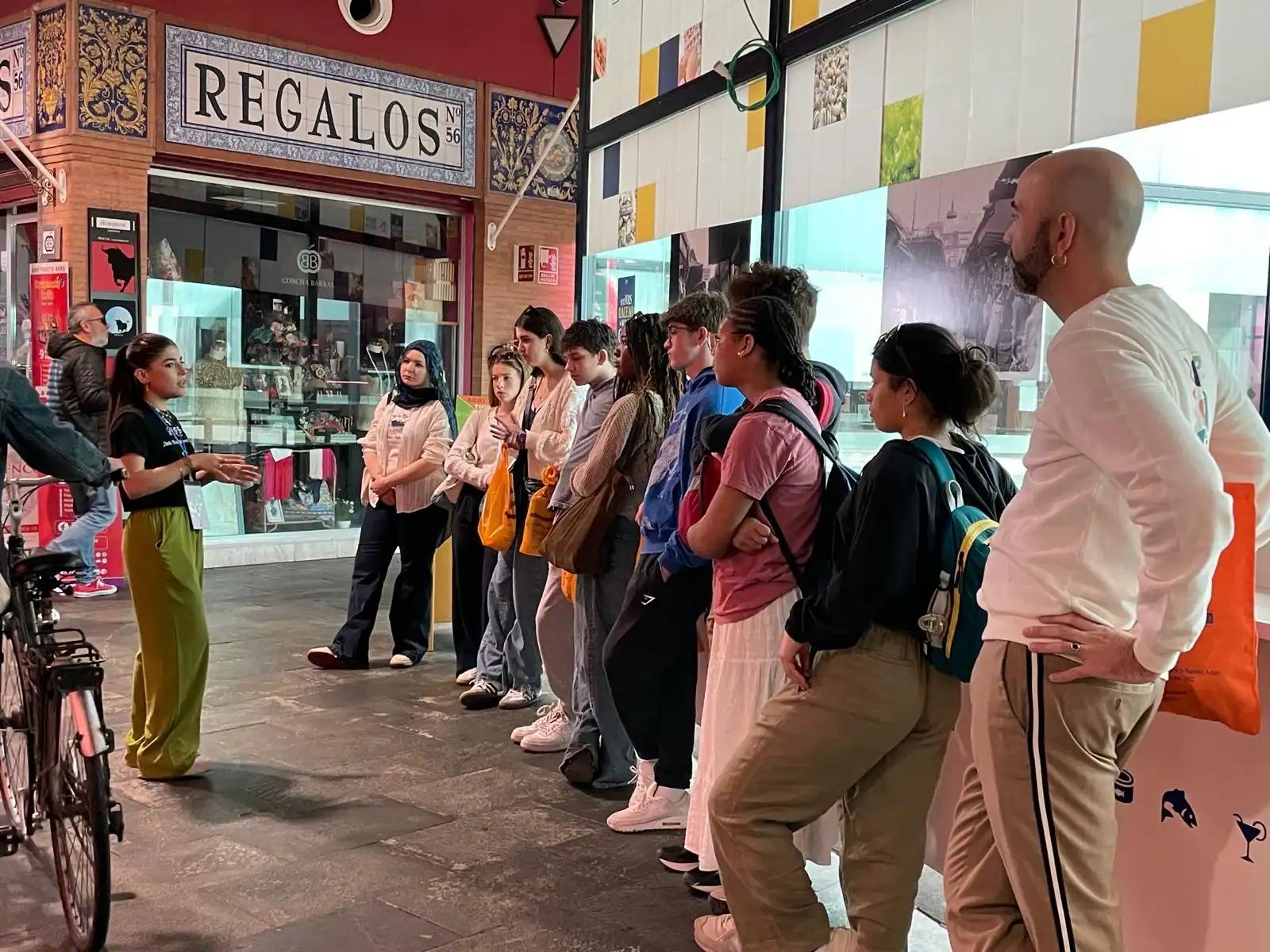 A group of people listens to a woman speaking inside a shop-lined hallway, with a "Regalos" sign visible above a storefront.