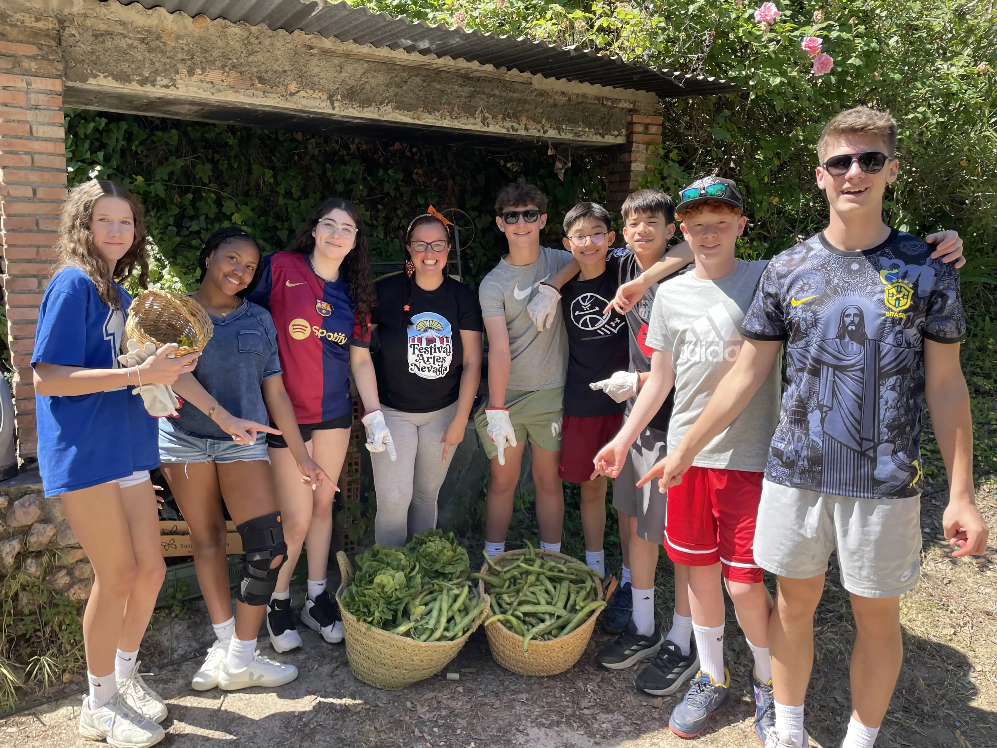 A group of eight teenagers stand outdoors, smiling by two large baskets filled with fresh green vegetables. Shrubs, a brick wall, and foliage are visible in the background.