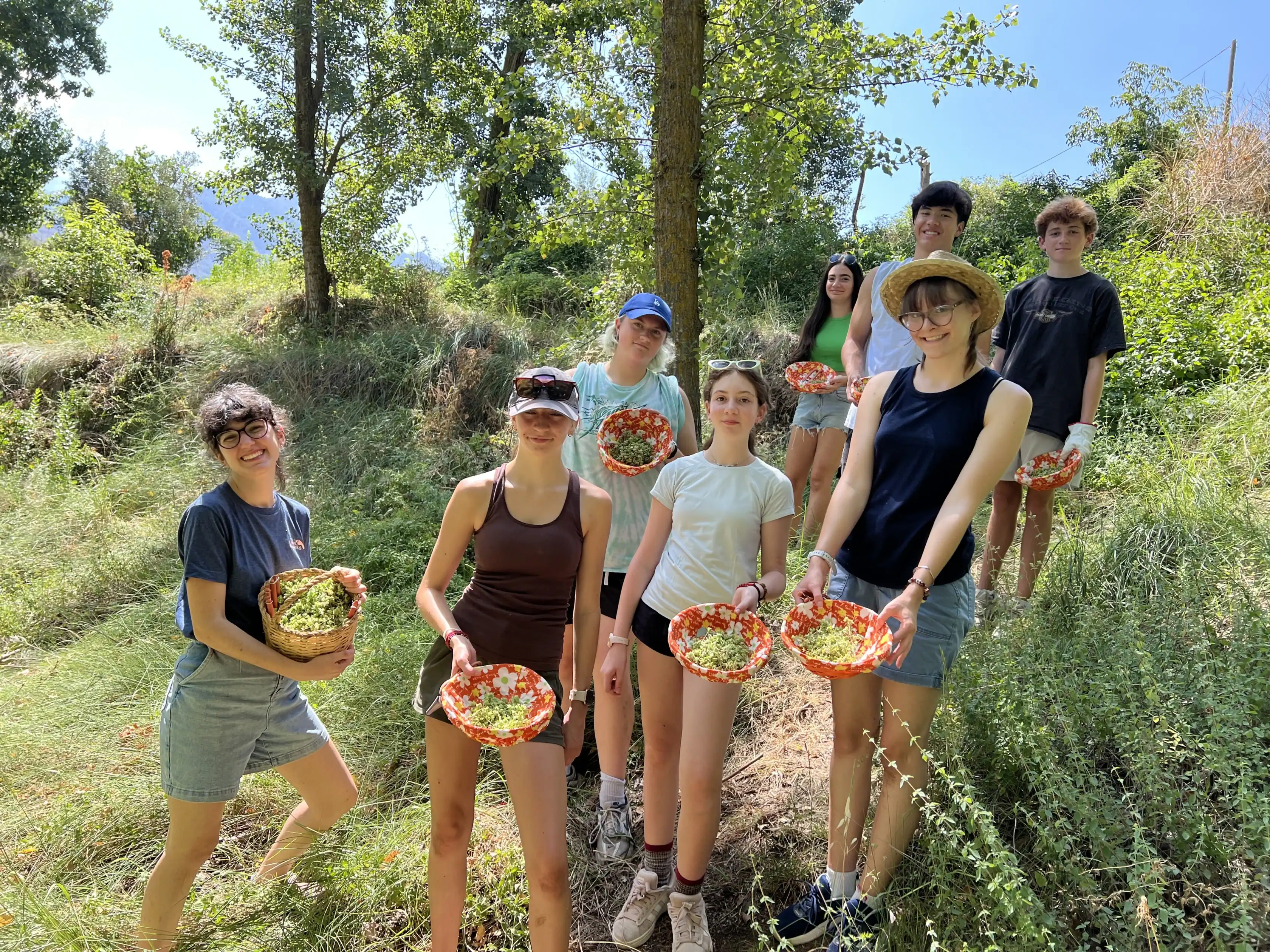 A group of eight young people stand outdoors on grassy terrain, each holding a basket filled with freshly picked produce, surrounded by trees and greenery.