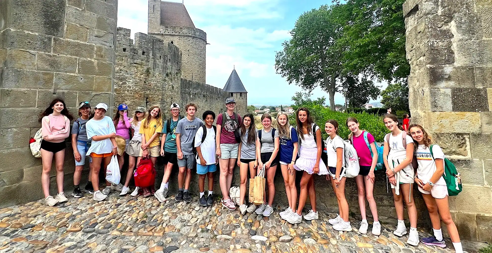 A group of teenagers poses together on a cobblestone path between stone castle walls, with towers and greenery in the background.