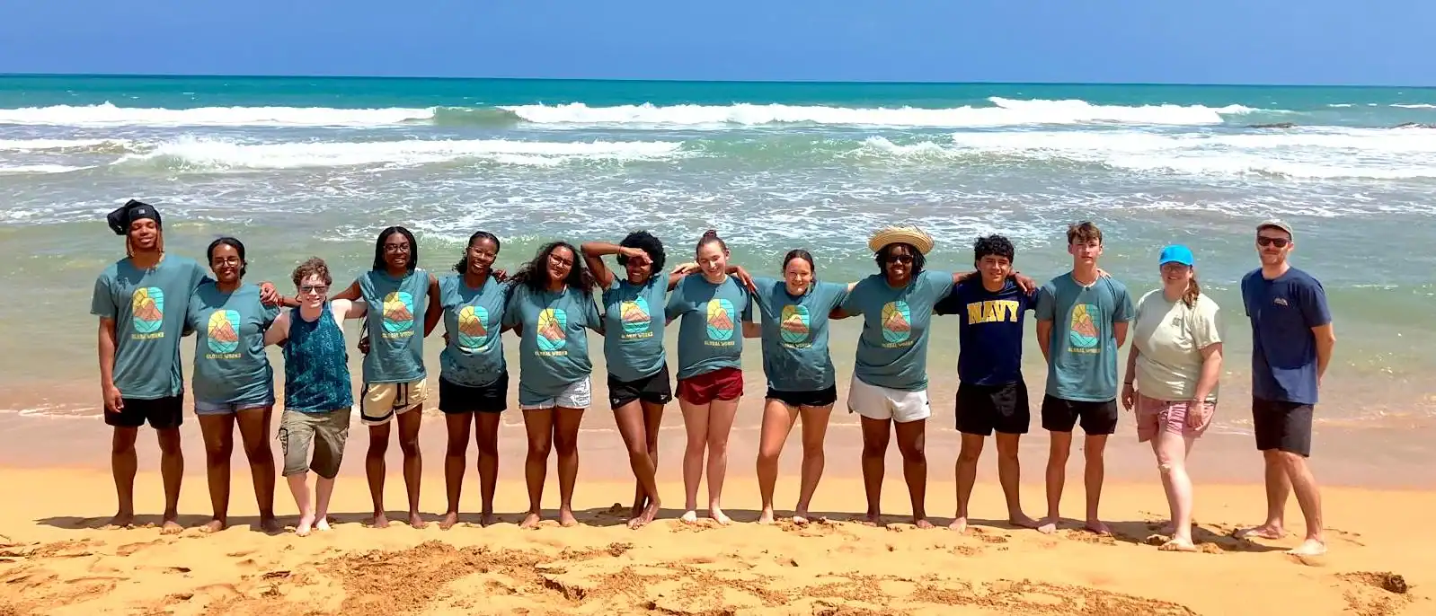 A group of thirteen people stand in a row on a sandy beach with ocean waves in the background, most wearing matching t-shirts.