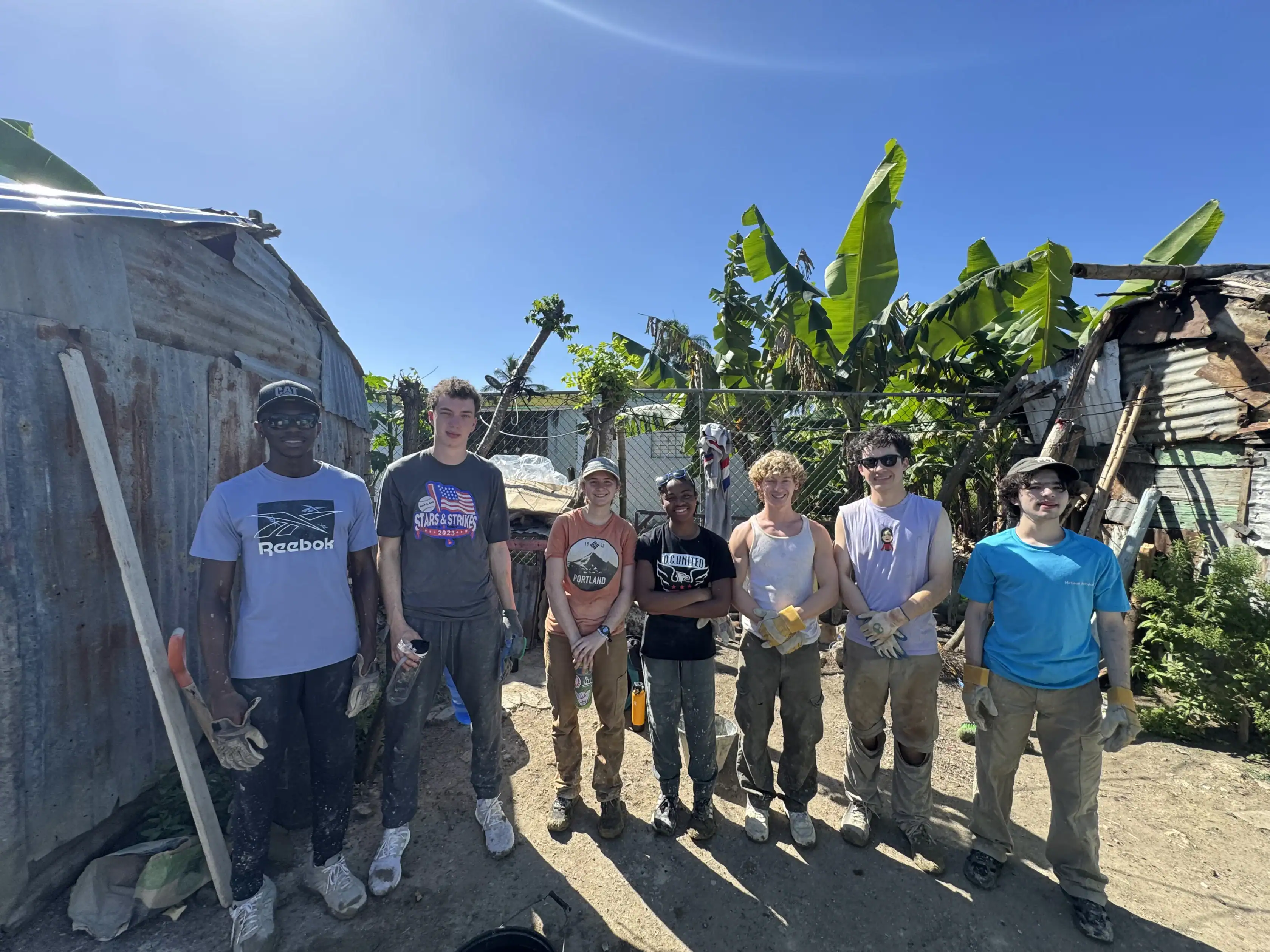 A group of seven people in work clothes and gloves stand outdoors in a line near rustic buildings and plants, posing for a photo on a sunny day.