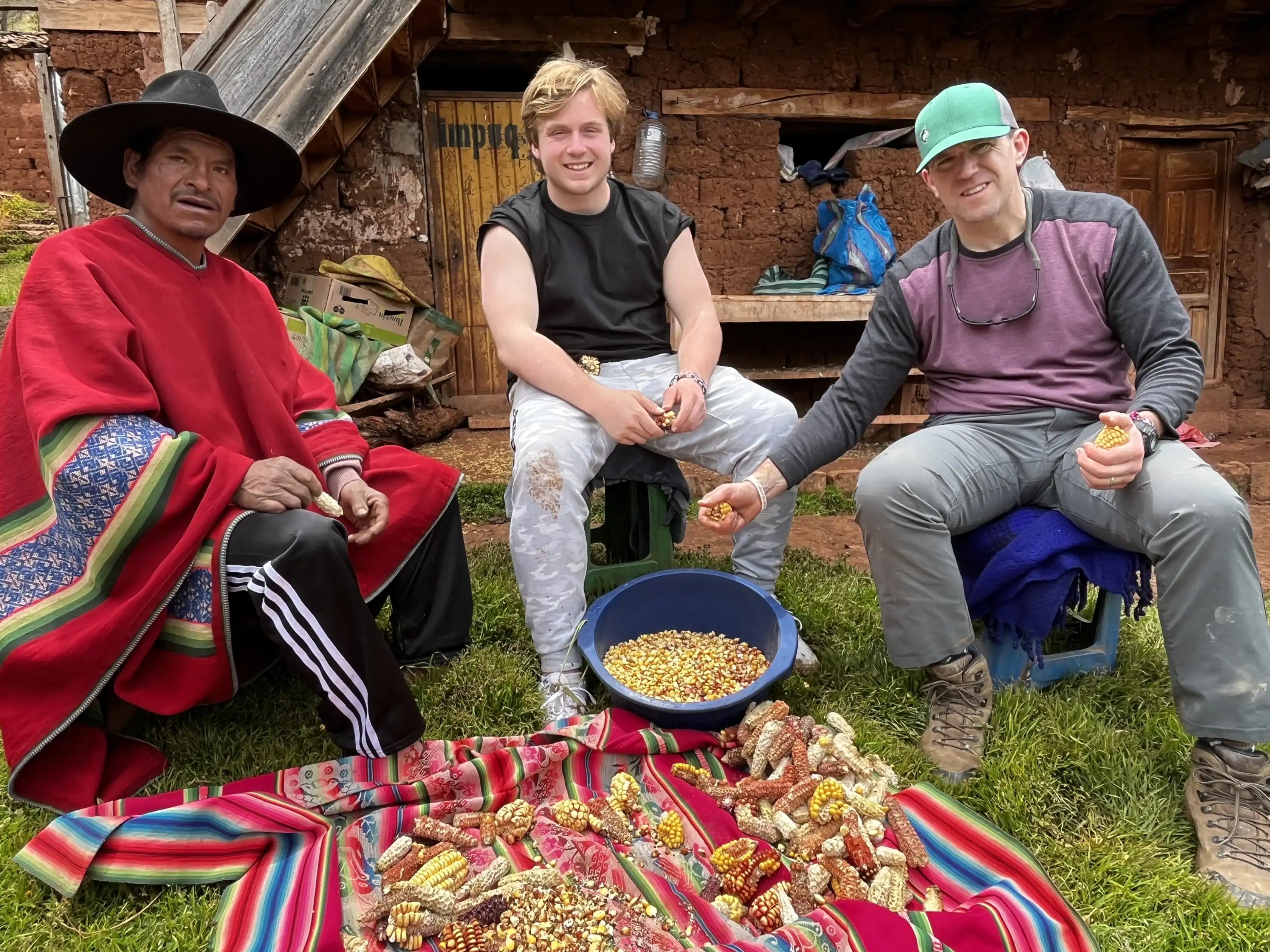 Three men sit outdoors, shelling corn cobs and collecting kernels in a blue bowl, with more colorful corn displayed on a red cloth in front of them.