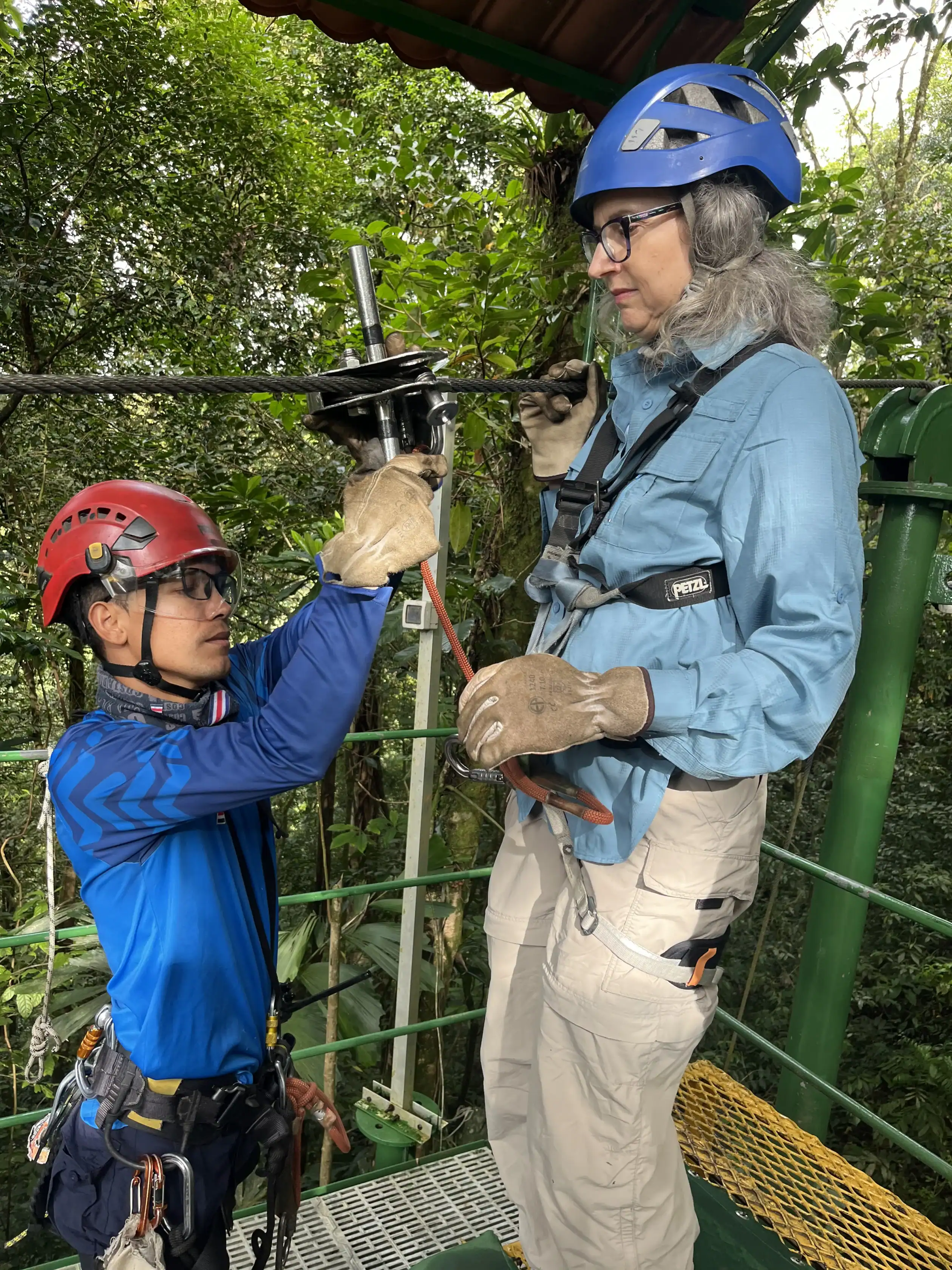 An instructor in blue assists a person with safety gear and helmet as they prepare to zipline in a forested area.