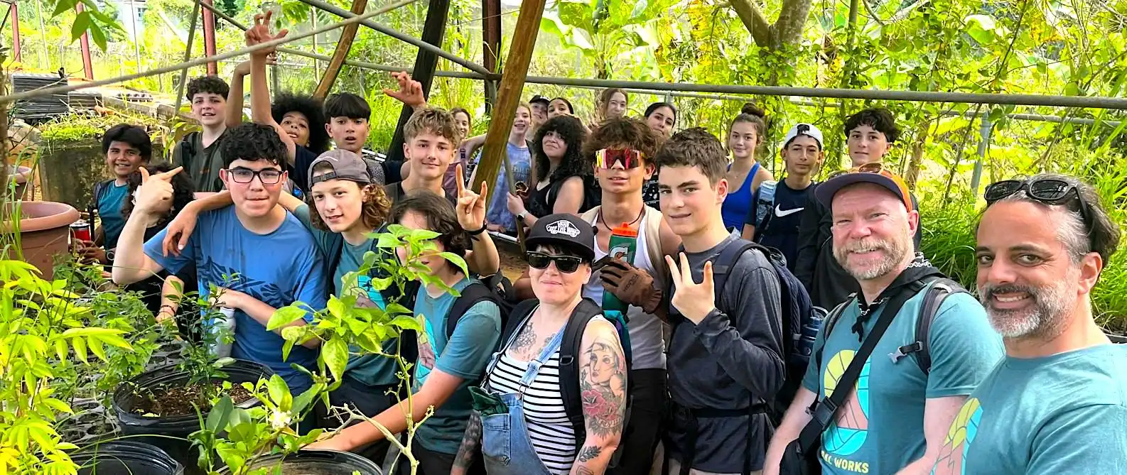 A group of teenagers and two adults pose together in a lush greenhouse, surrounded by green plants and potted seedlings.