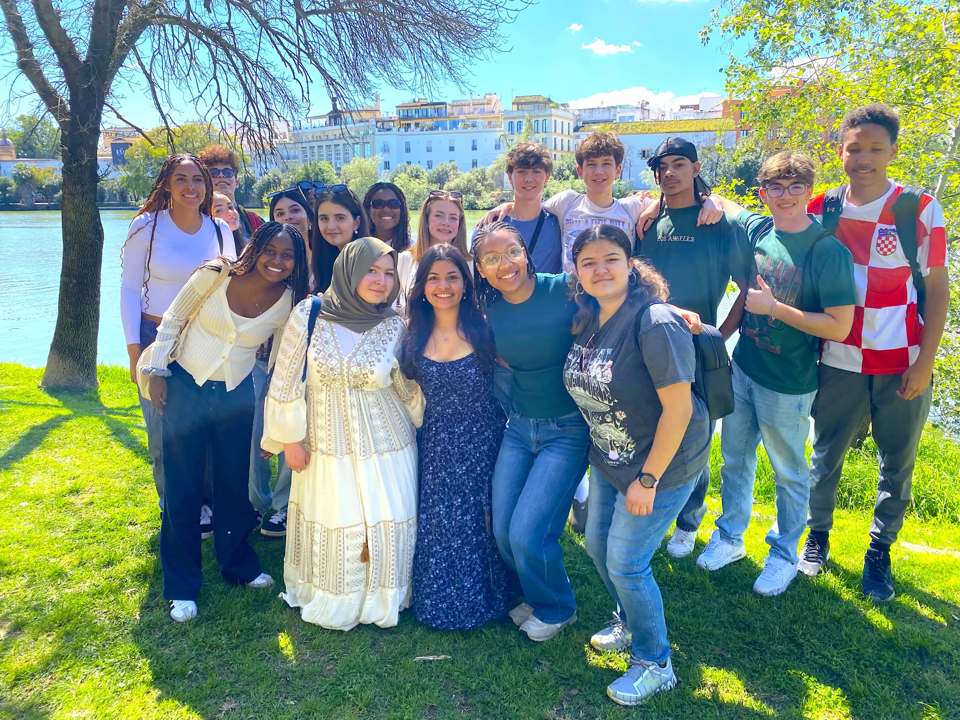 A group of sixteen teenagers poses together on grass near a river, with buildings and trees in the background on a sunny day.