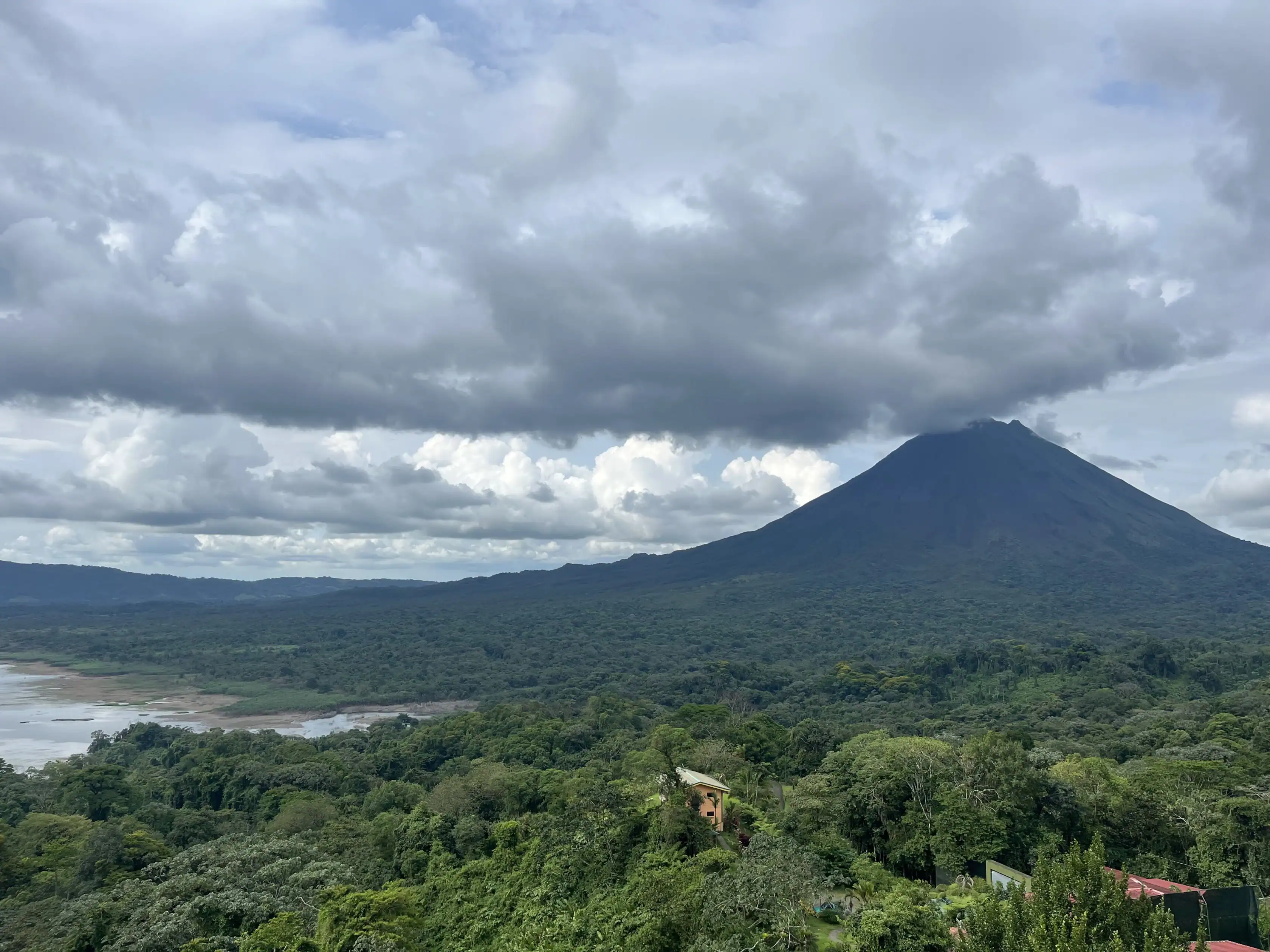 A large volcanic mountain rises above a dense green forest under a cloudy sky, with a body of water visible on the left side of the landscape.