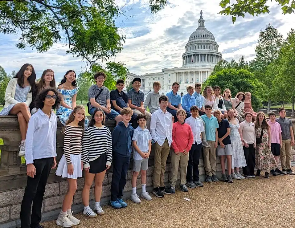 A group of students and adults pose outdoors in front of the U.S. Capitol building on a cloudy day, surrounded by trees and greenery.