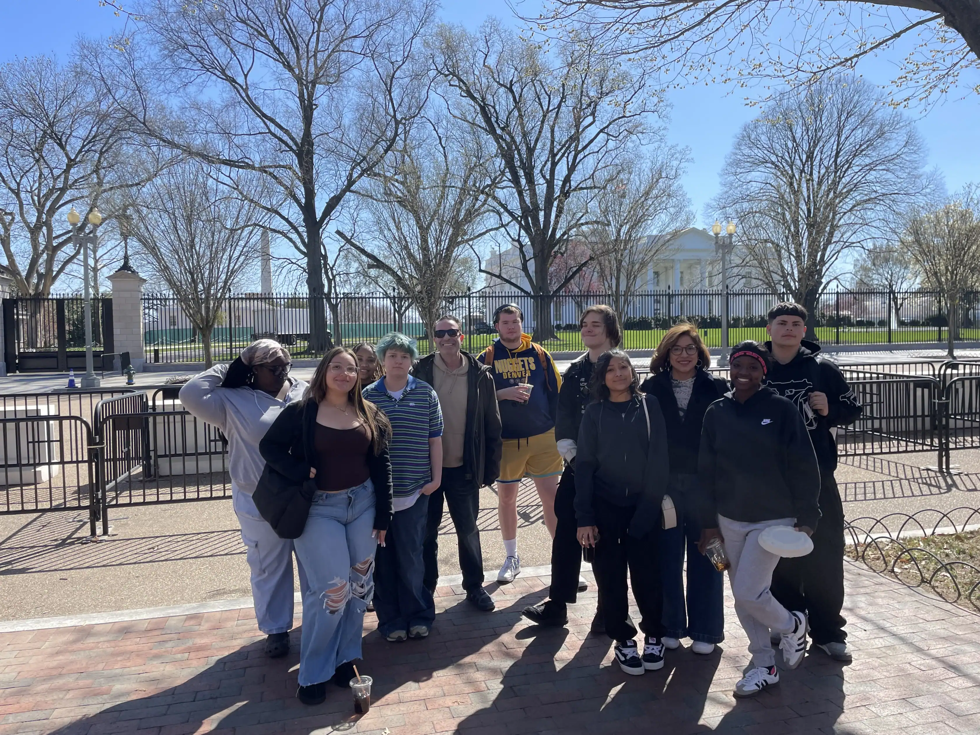 A group of people pose for a photo outside on a sunny day in front of the White House, with bare trees and a metal fence in the background.