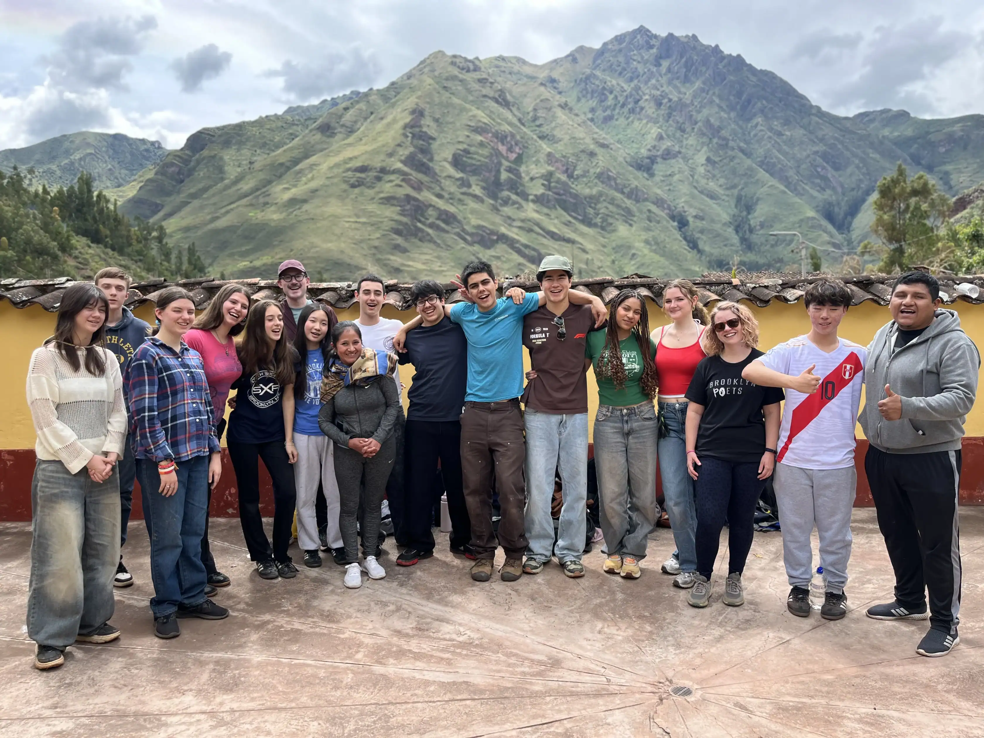 A group of students pose together on a school trip on a patio with green mountains and cloudy sky in the background.