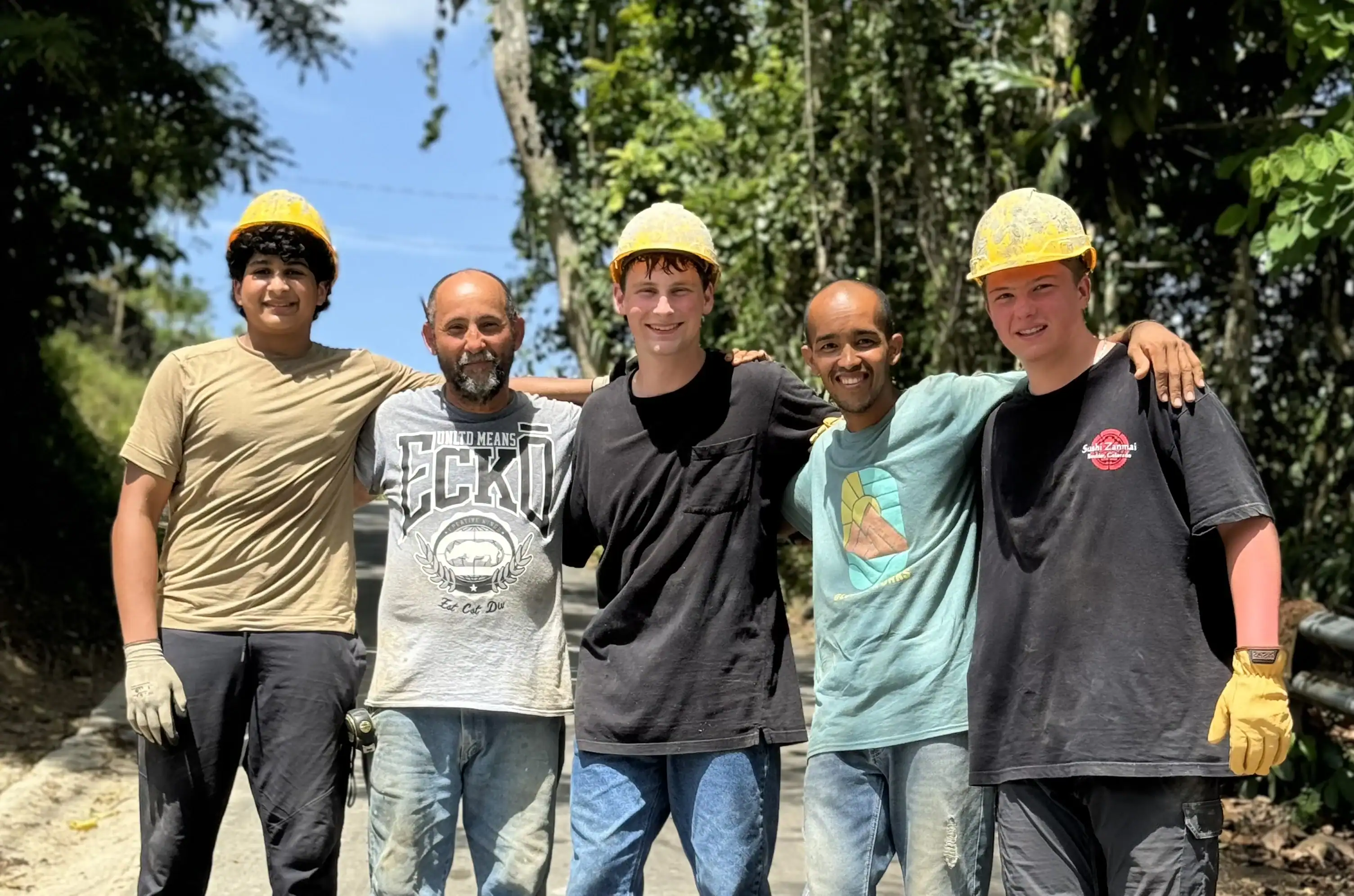 Five people wearing work gloves and yellow safety helmets stand together outdoors on a sunny day, smiling with their arms around each other. Trees and foliage are visible in the background.