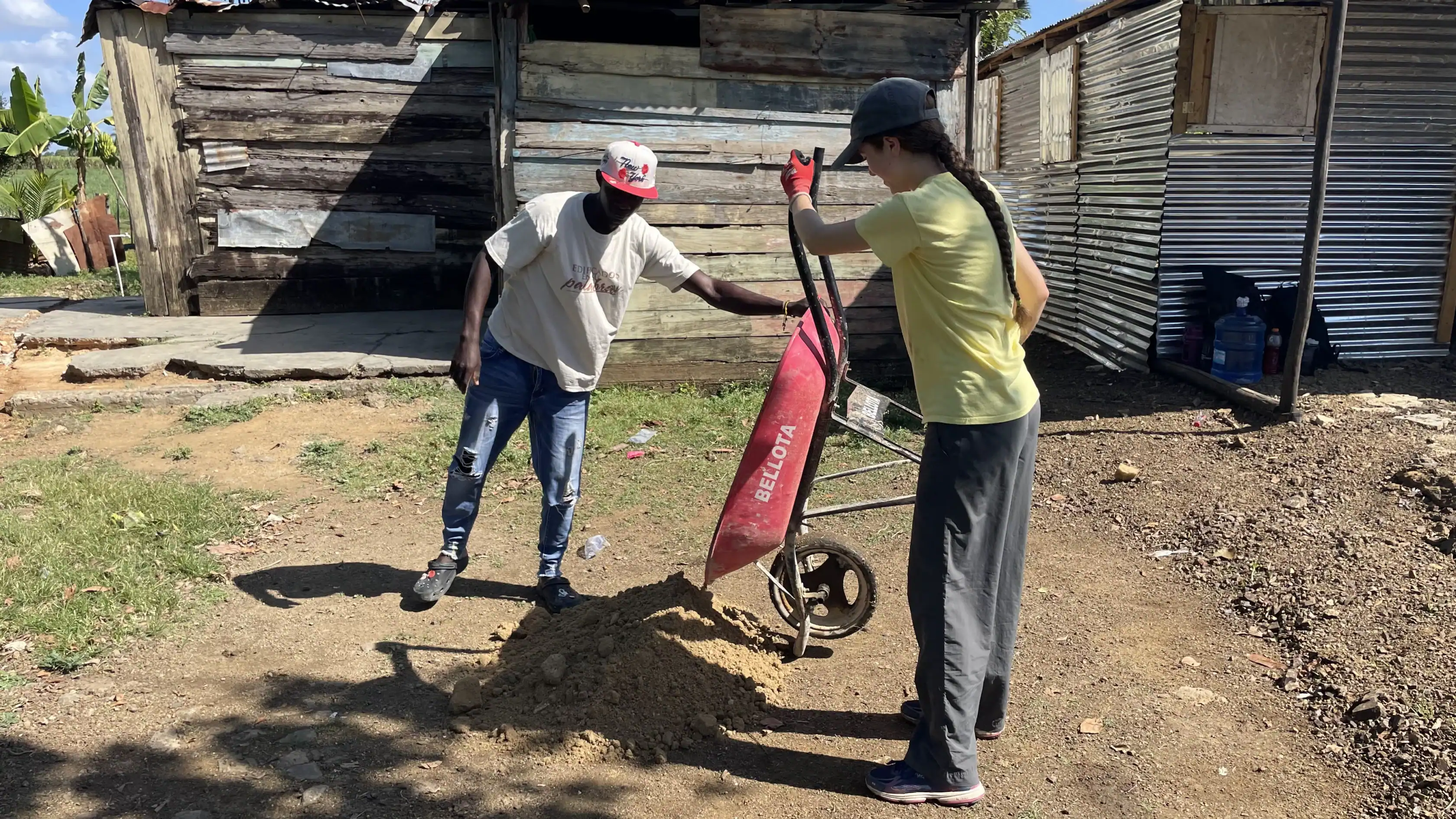 Two people work outdoors near wooden buildings; one holds a wheelbarrow over a pile of dirt while the other points at the ground.