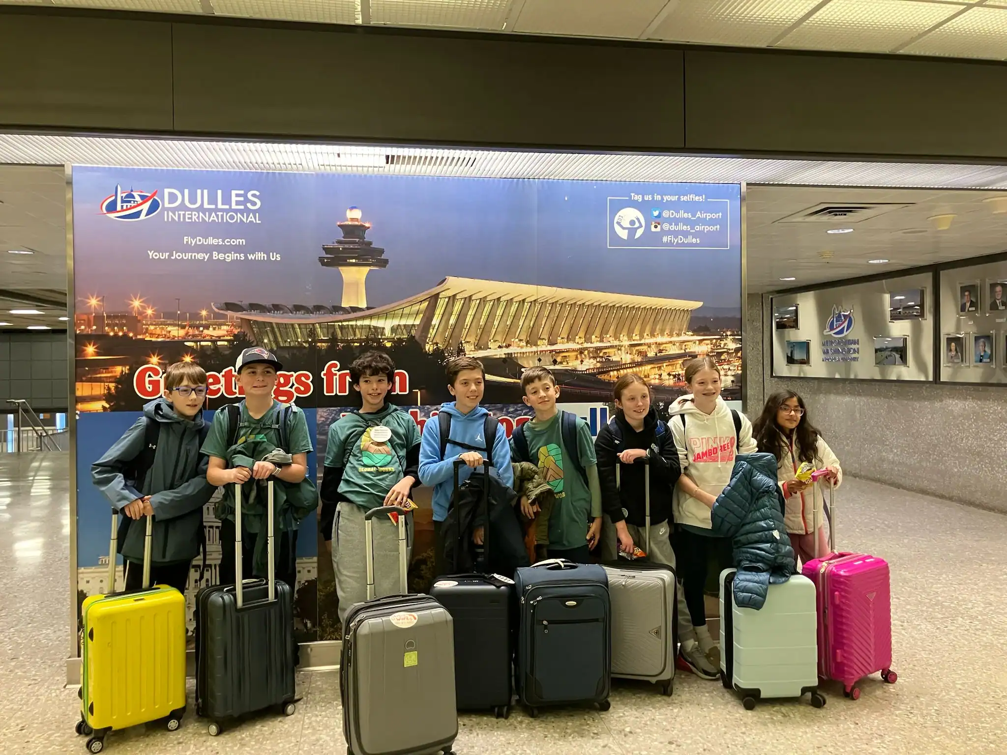 Seven children stand with suitcases in front of a sign for Dulles International Airport inside a terminal.