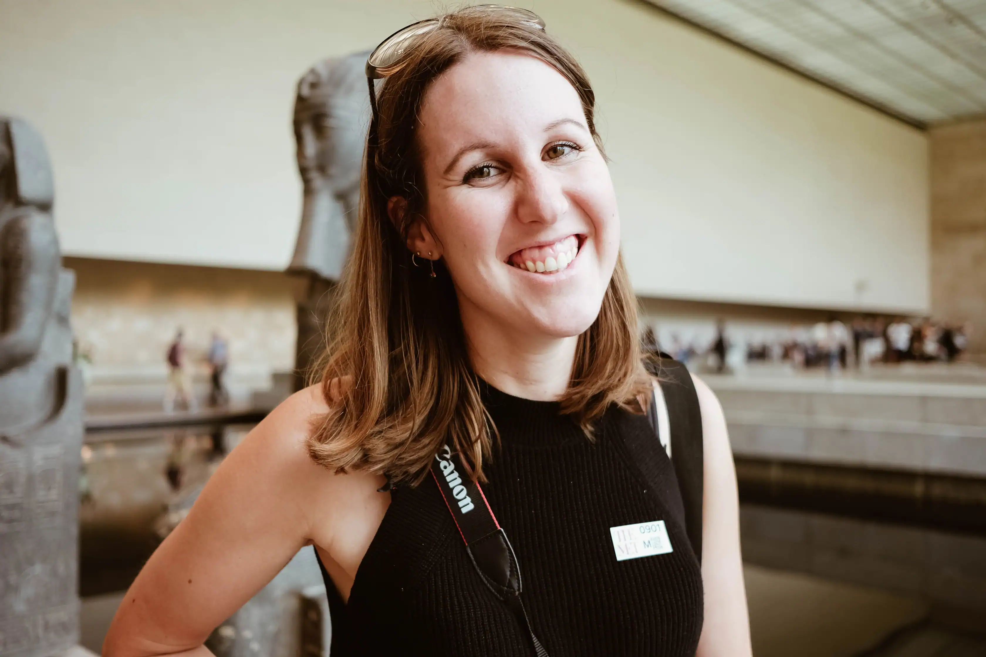 A woman with shoulder-length brown hair smiles at the camera inside a museum, wearing a black sleeveless top, a name tag, and a camera strap around her neck.