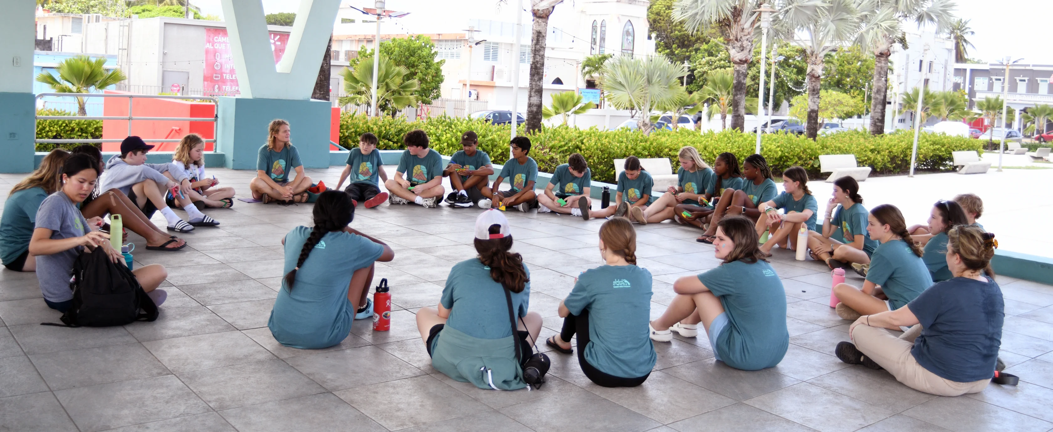 A group of people in matching blue shirts sit in a large circle on a tiled floor outdoors, engaged in a group activity or discussion.