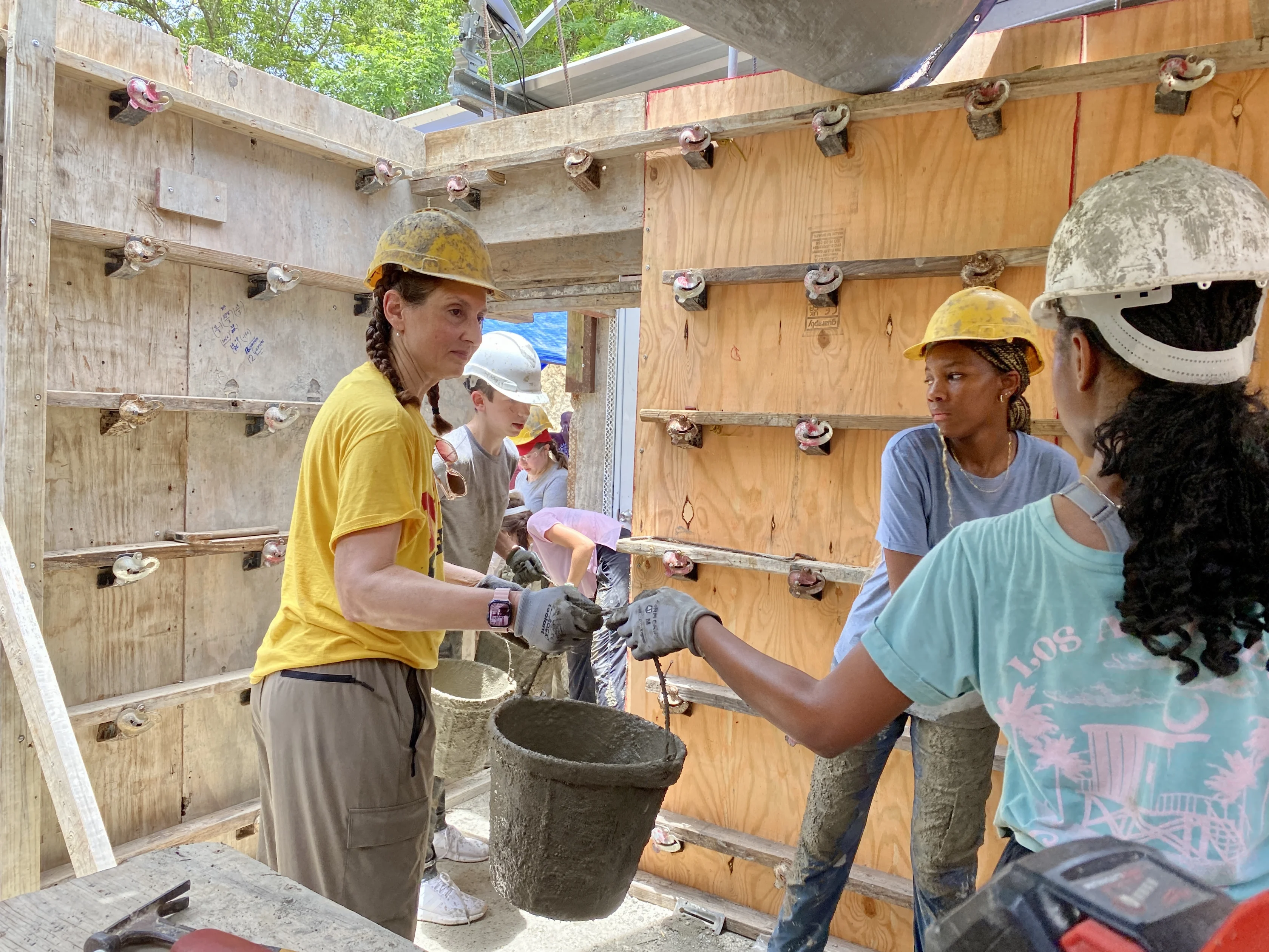 Several people wearing hard hats and gloves work together to pass buckets of concrete at a construction site with wooden formwork in the background.