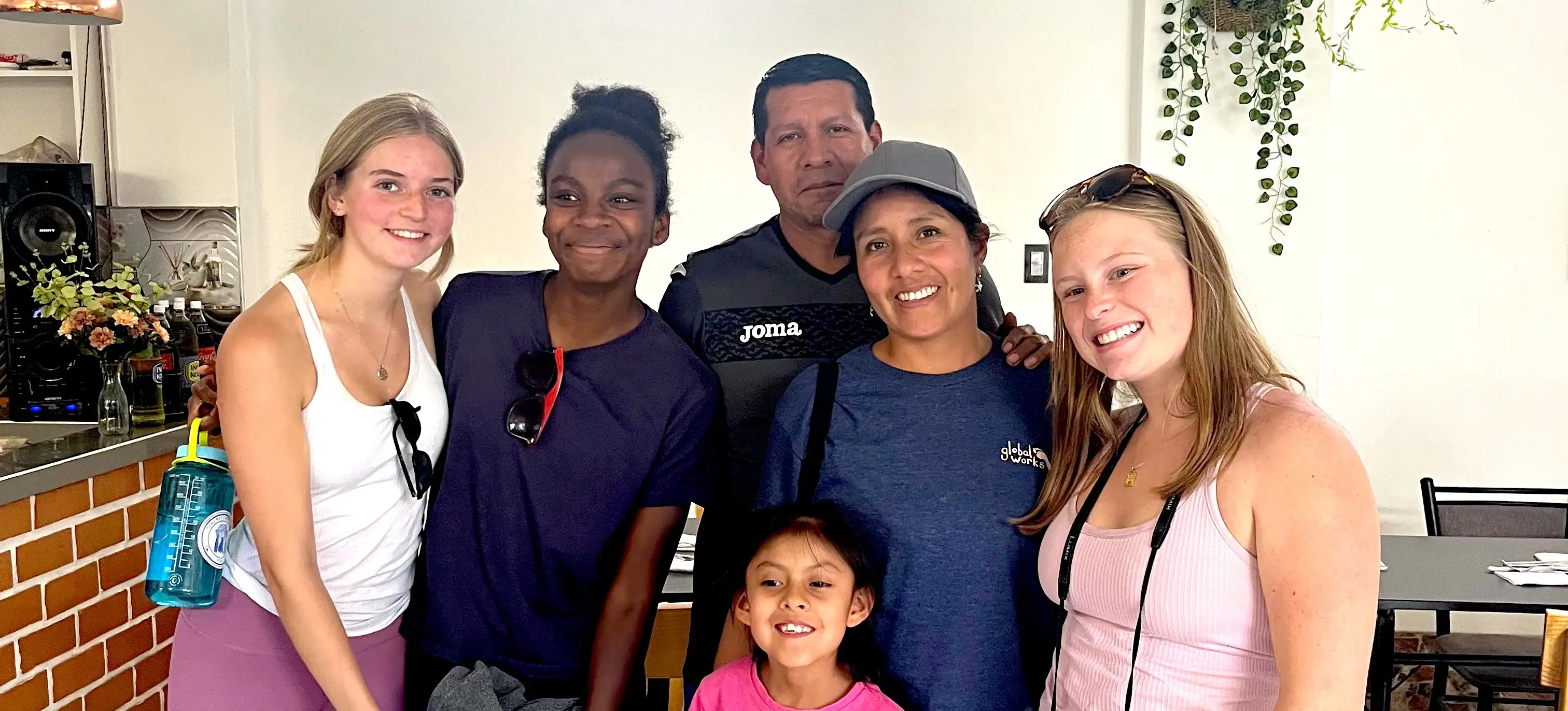 A group of five adults and one child stand close together indoors, smiling at the camera. There are plants and a brick counter in the background.
