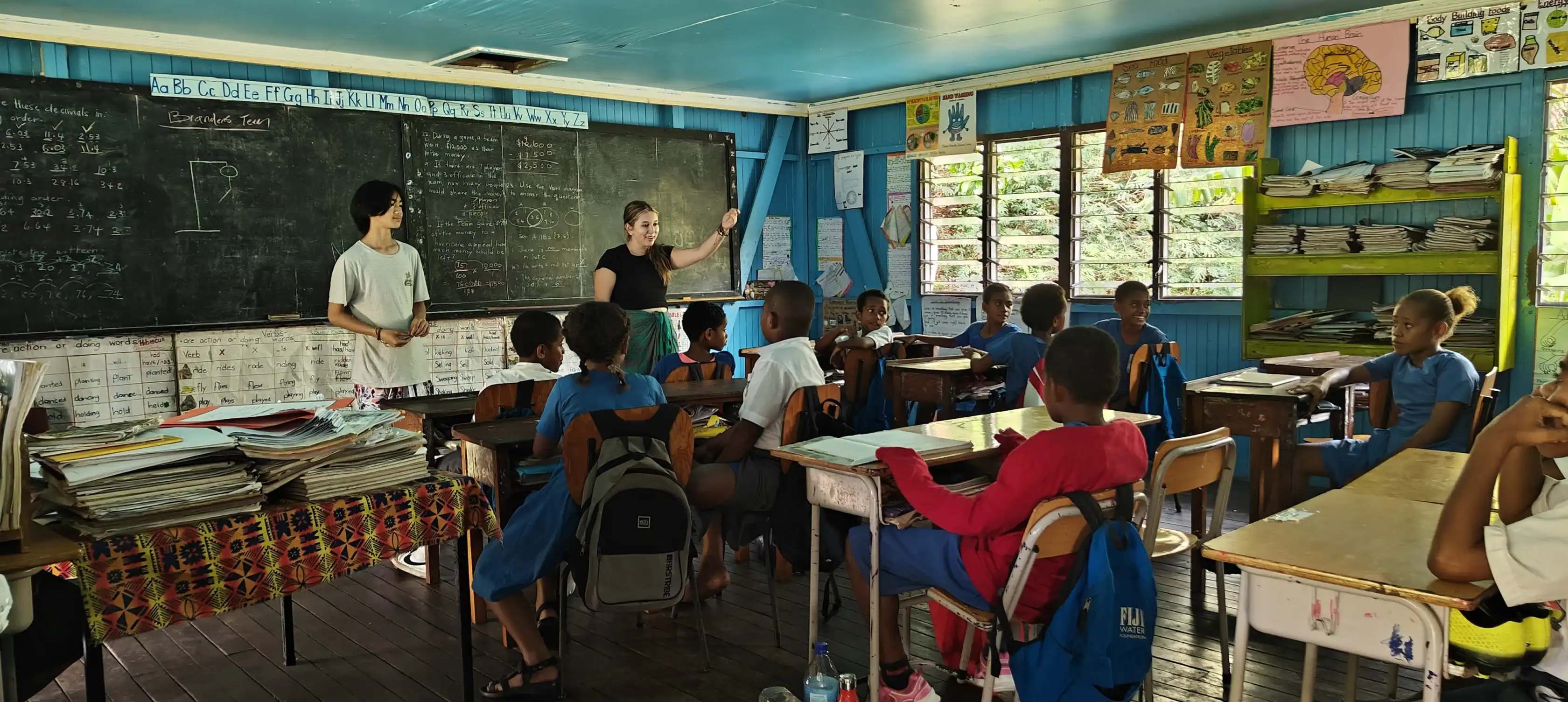 Two teachers stand at the front of a classroom, instructing a group of seated students who are facing the chalkboard in a brightly lit, colorful classroom.