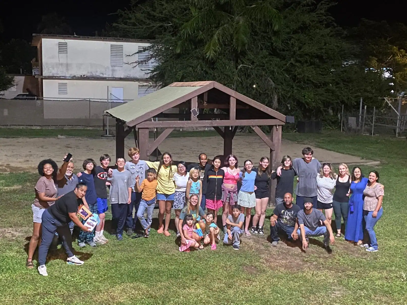 A large group of people, including children and adults, pose together at night under a wooden shelter on a grassy area.