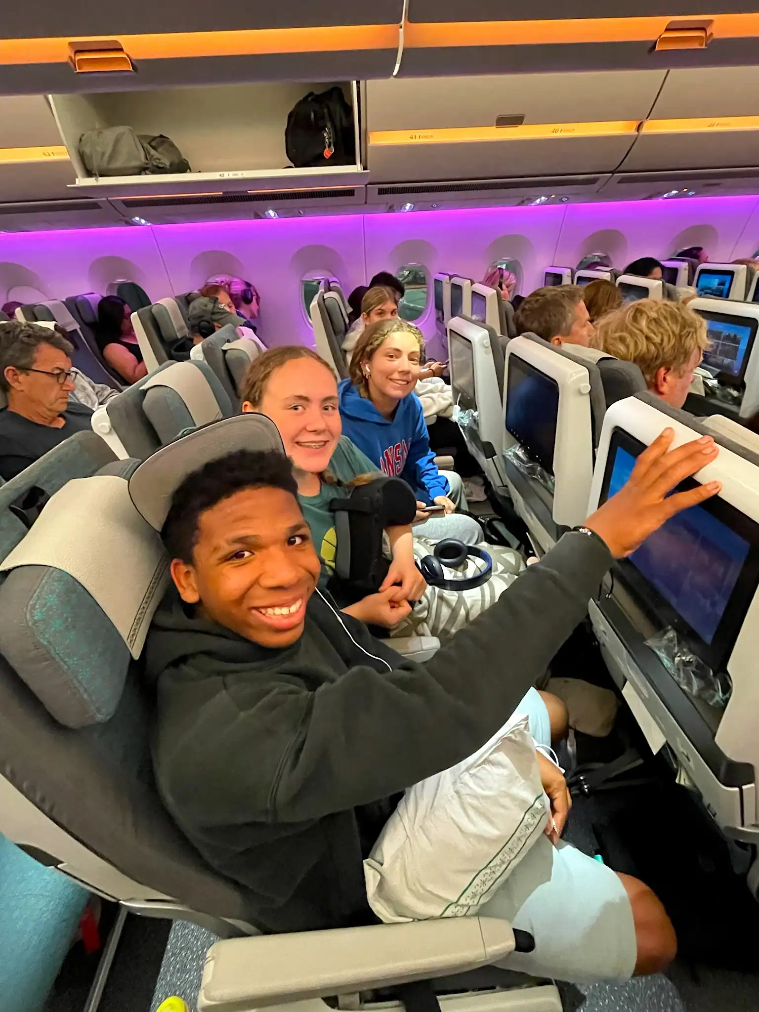 A group of passengers sitting in economy seats on an airplane, some smiling at the camera, with in-flight screens visible.