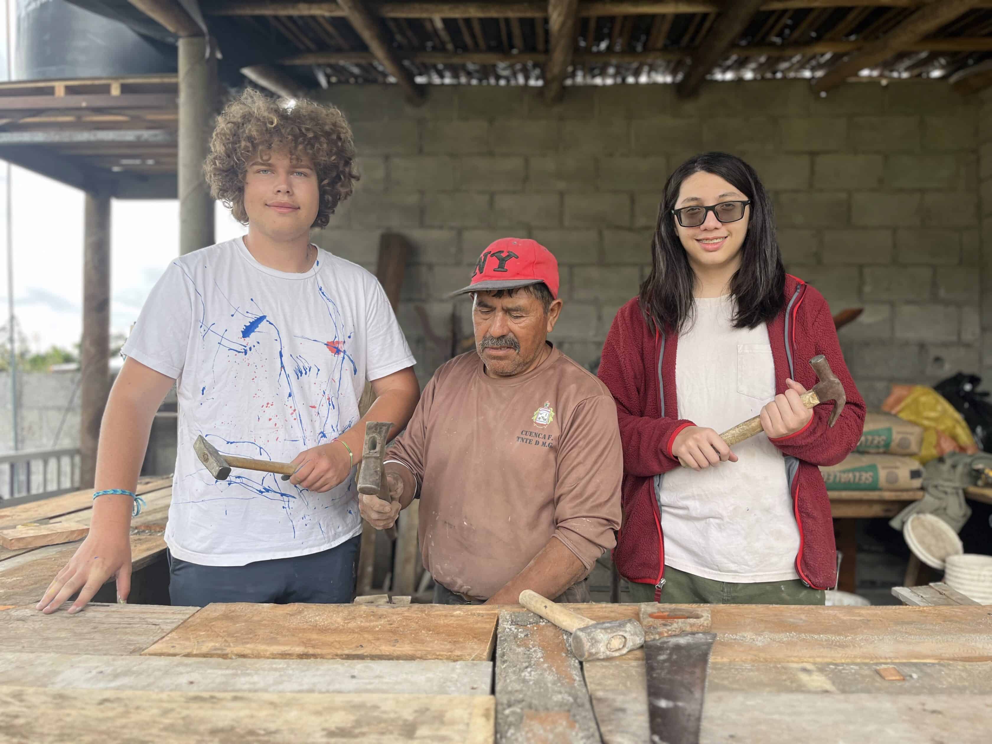 Two teenagers and an older man stand at a wooden workbench holding carpentry tools, with wooden planks and building materials in the background, embodying the spirit of teamwork found on teen service trips.