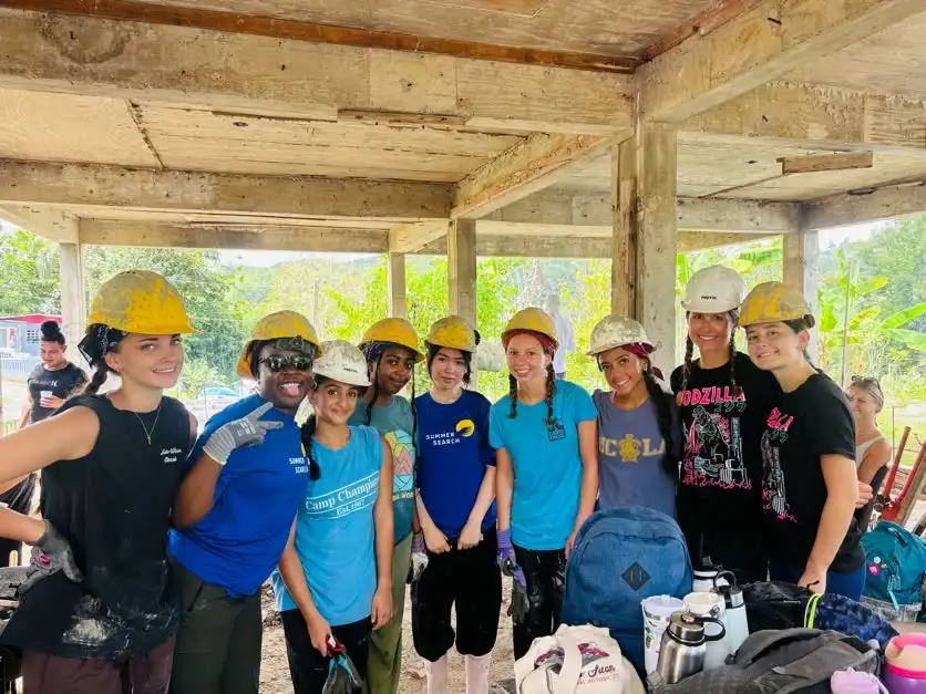 A group of nine people wearing yellow and white hard hats pose together under a concrete structure at a construction site, surrounded by bags and tools—capturing the teamwork spirit often found on teen service trips.