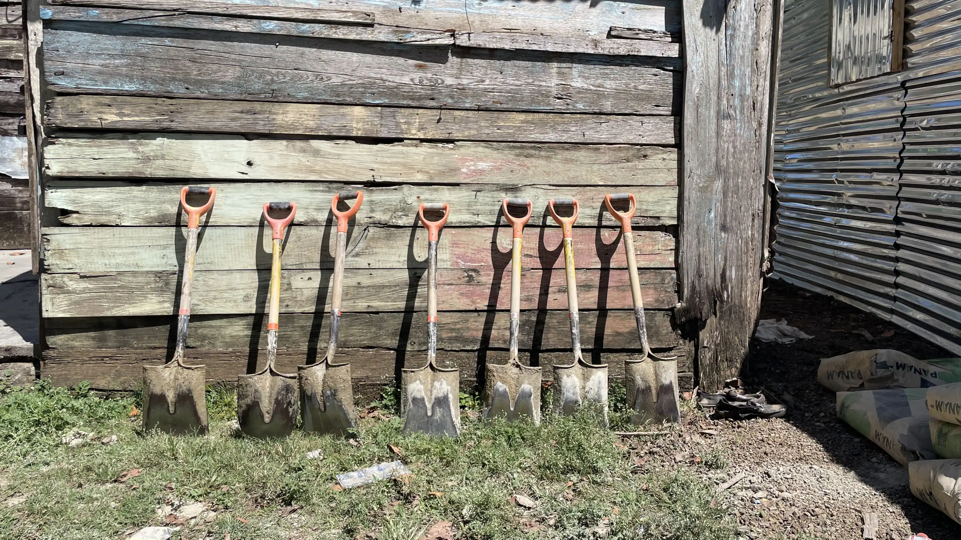 Eight shovels with orange handles are lined up against a weathered wooden wall outdoors, ready for action on teen service trips, with grass and dirt in the foreground.