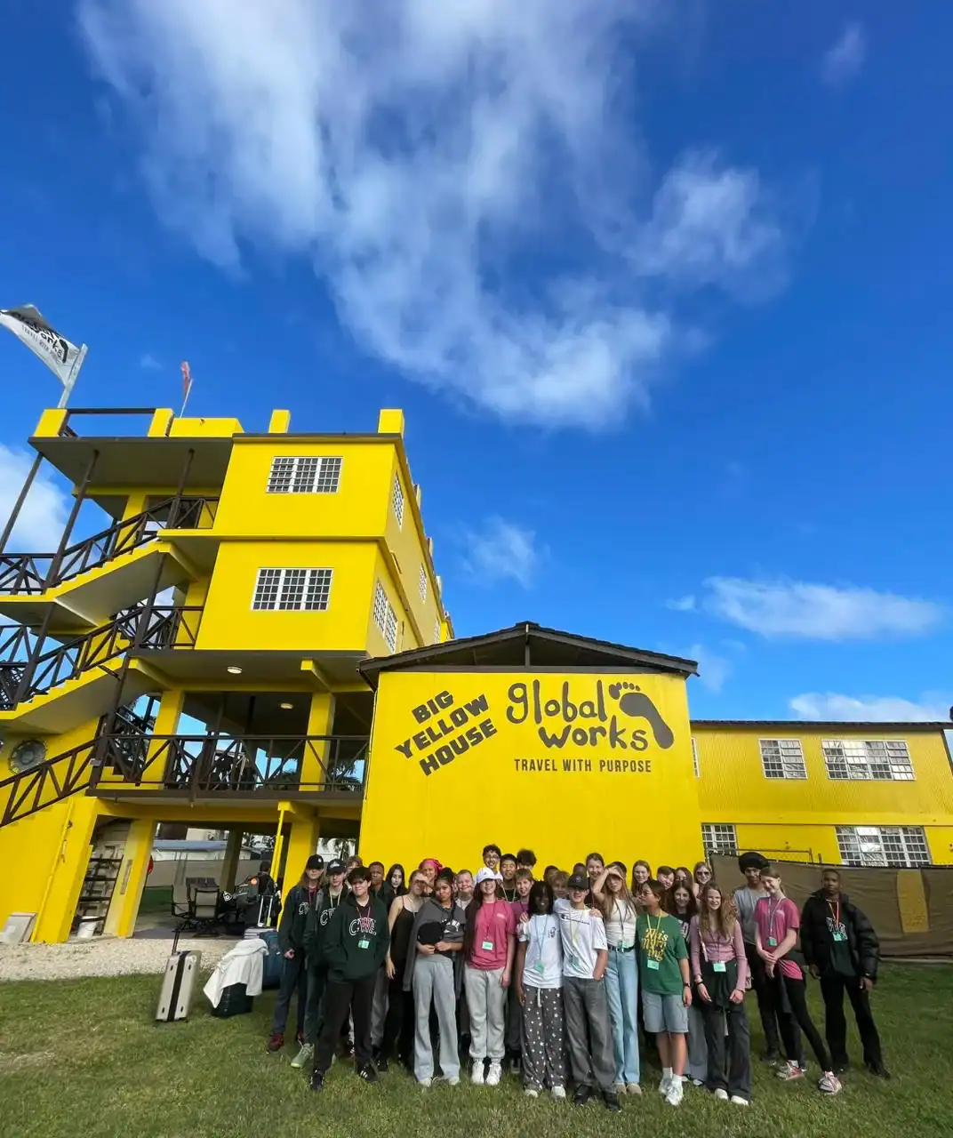 A large group of people stands in front of a bright yellow building labeled "Big Yellow House, Global Works, Travel With Purpose" under a blue sky with scattered clouds.