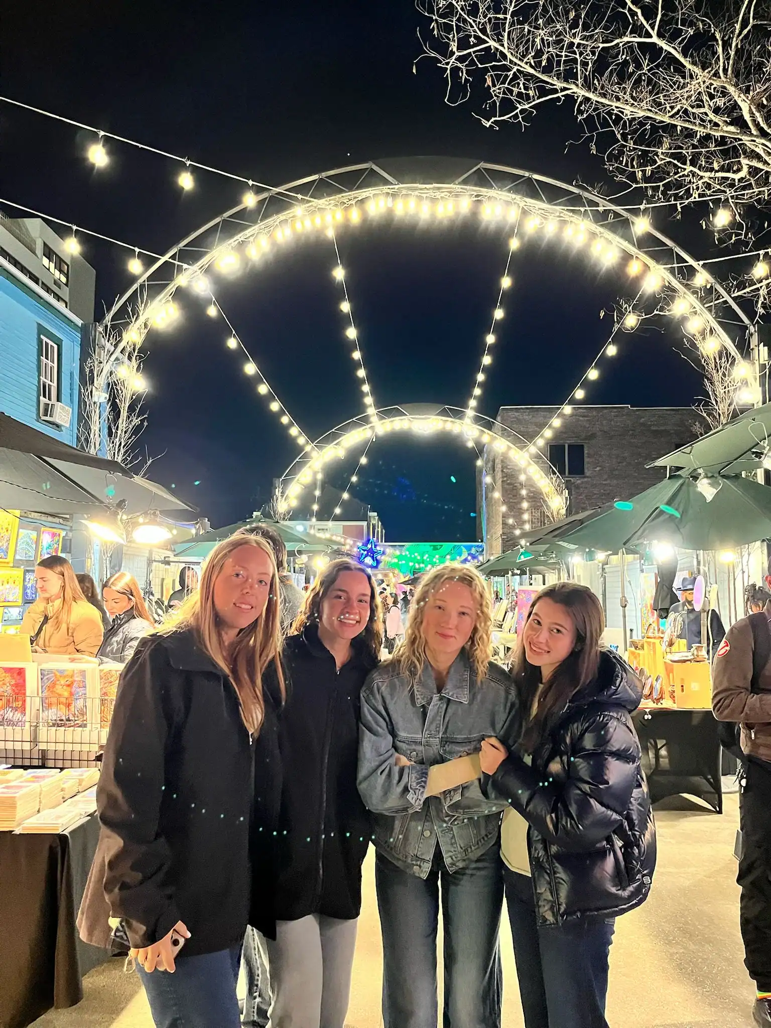 Four women stand together at an outdoor night market under arching string lights, surrounded by vendor booths and illuminated artwork displays.