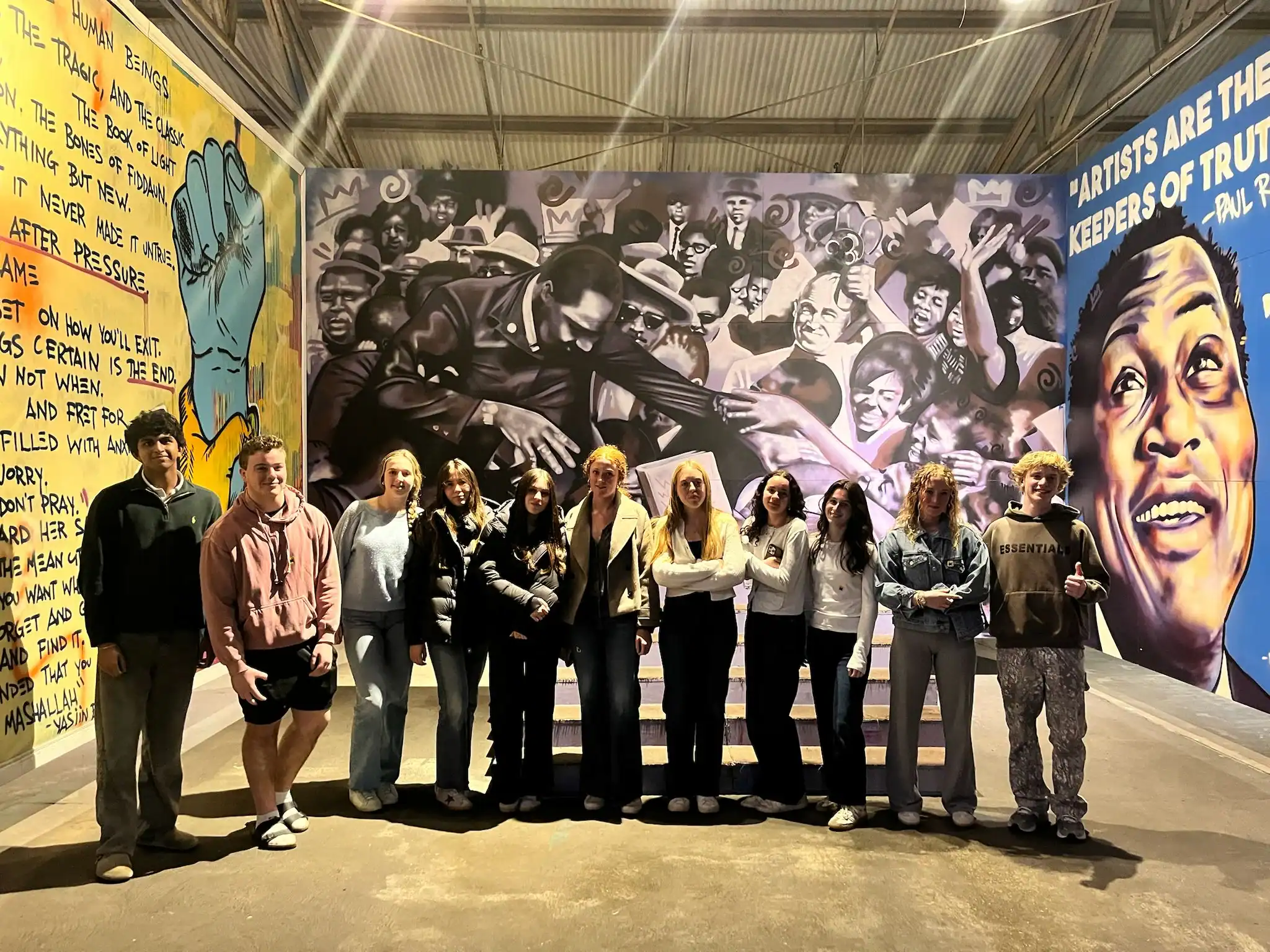 A group of eleven people stands side by side in front of a colorful mural featuring civil rights imagery and prominent figures inside an art gallery.